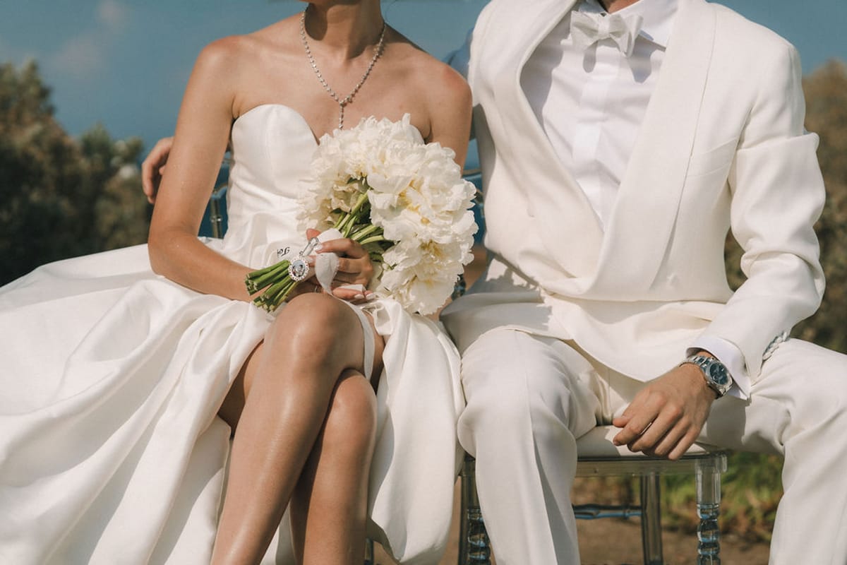 The couple sit side by side in white, the bride holding a bouquet of white flowers on her lap.