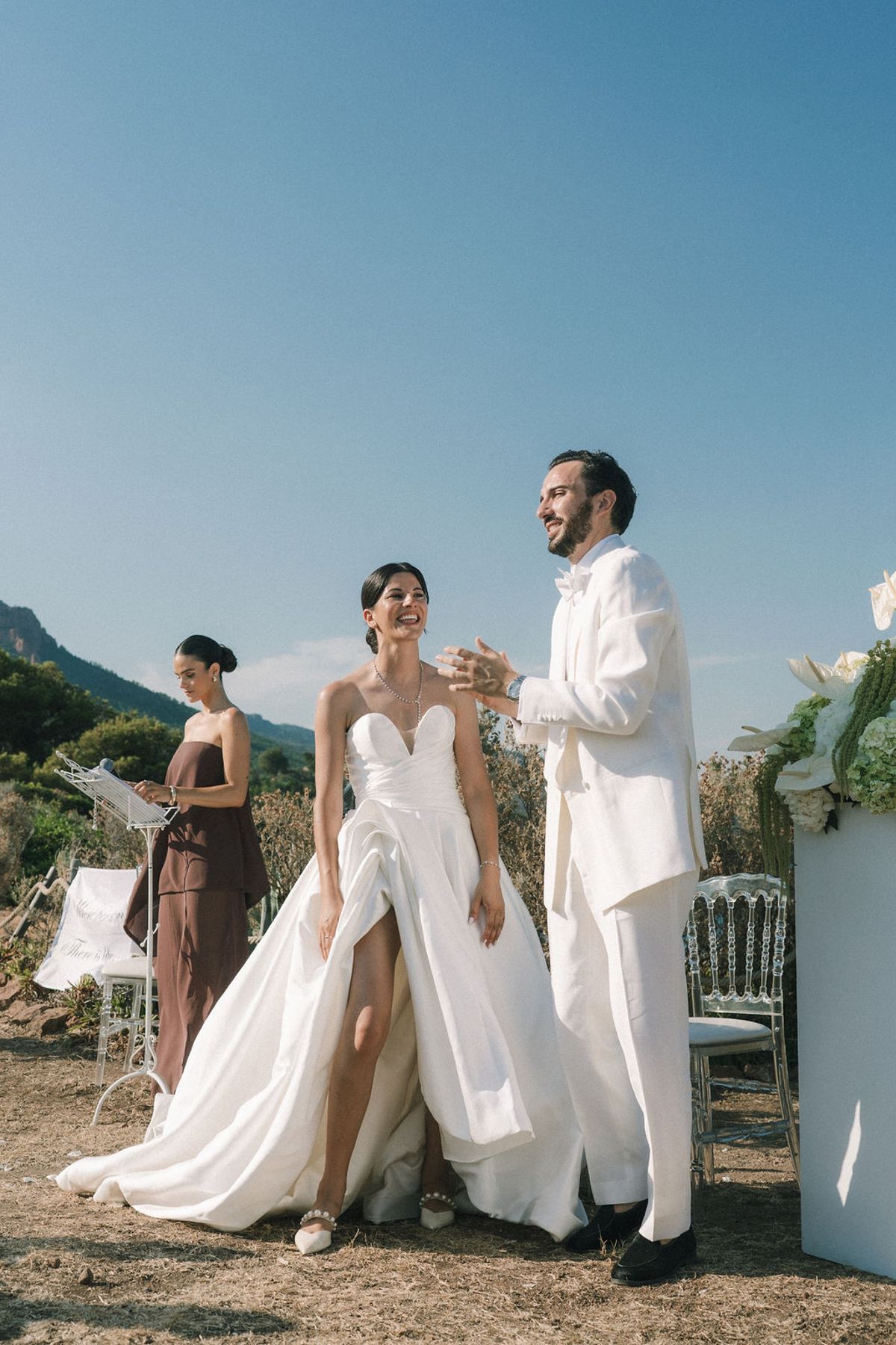 The bride in a white gown and the groom in a white suit stand smiling beside an officiant on a sunny coastal hillside.