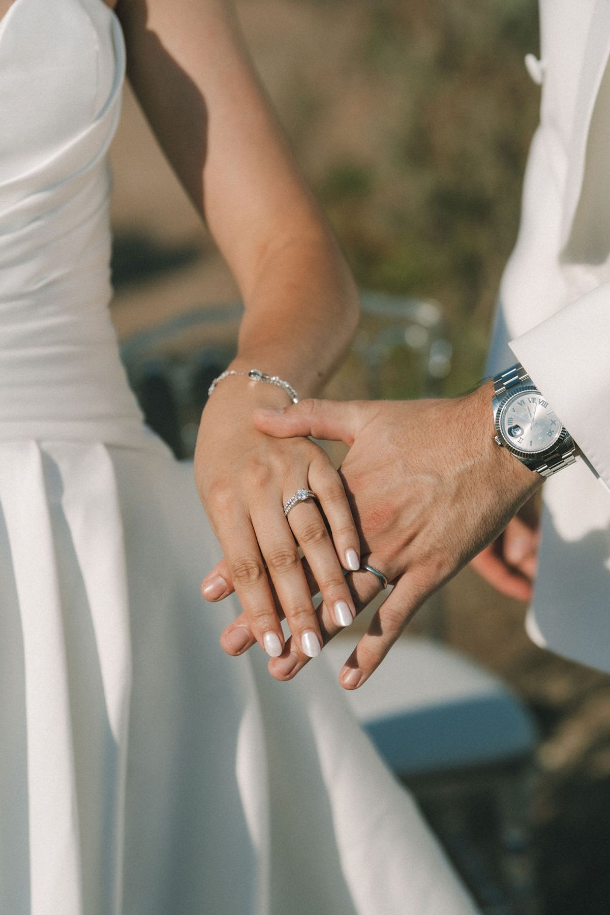 The couple's hands rest together, showing rings and a wristwatch, beside the bride's white skirt and the groom's white jacket.