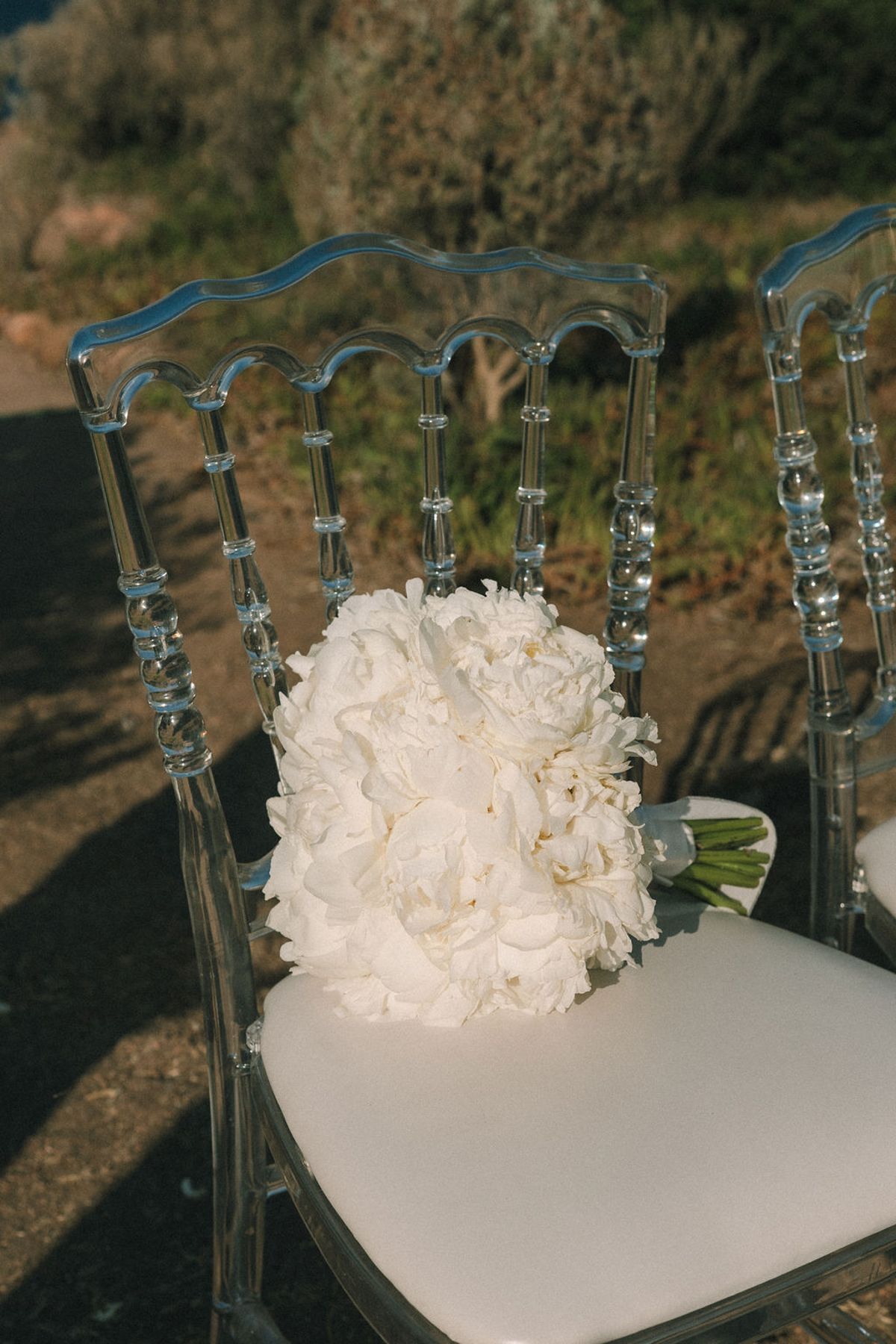 A large bunch of white flowers rests on the seat of a transparent chair with a white cushion on dry ground.