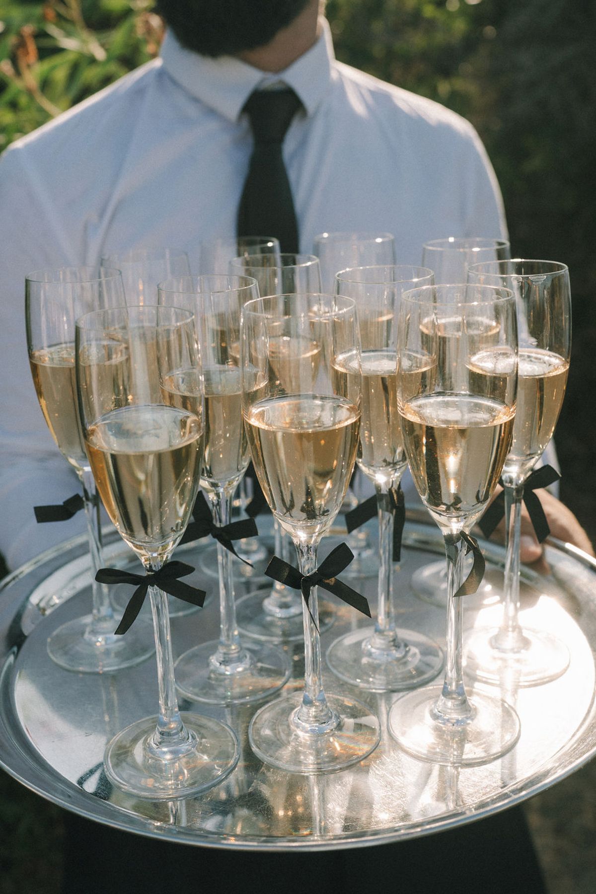 A person in a white shirt and black tie holds a silver tray of champagne flutes tied with black ribbons.