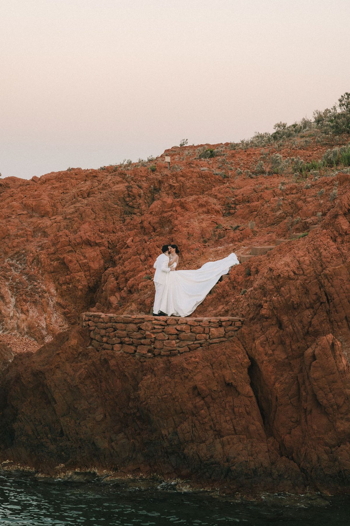 The couple embrace on a ledge of red rock above the water, the bride's white dress blowing in the wind.