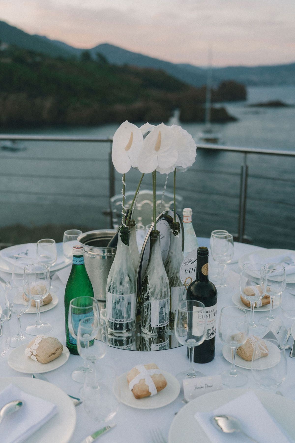 A round table set with plates, bread, glasses and bottles around a white flower centrepiece, with a bay and hills behind.