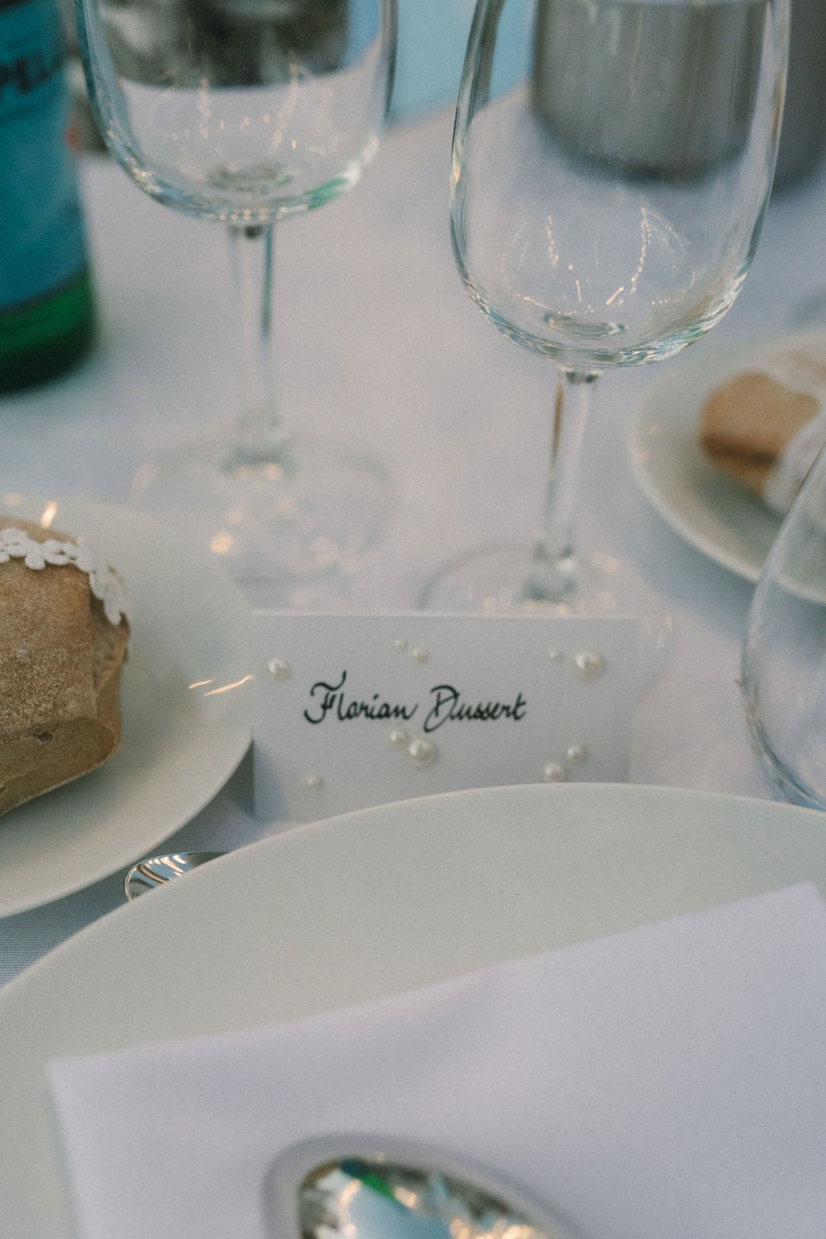 A white tablecloth holds wine glasses, a bread roll, a plate and a small handwritten name card with pearl details.