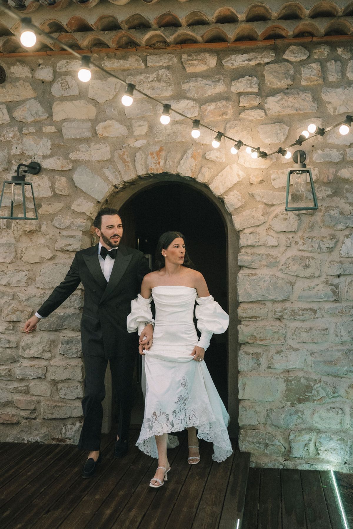 The couple walk hand in hand out of an arched stone doorway beneath strings of lights, the groom in a dark suit.