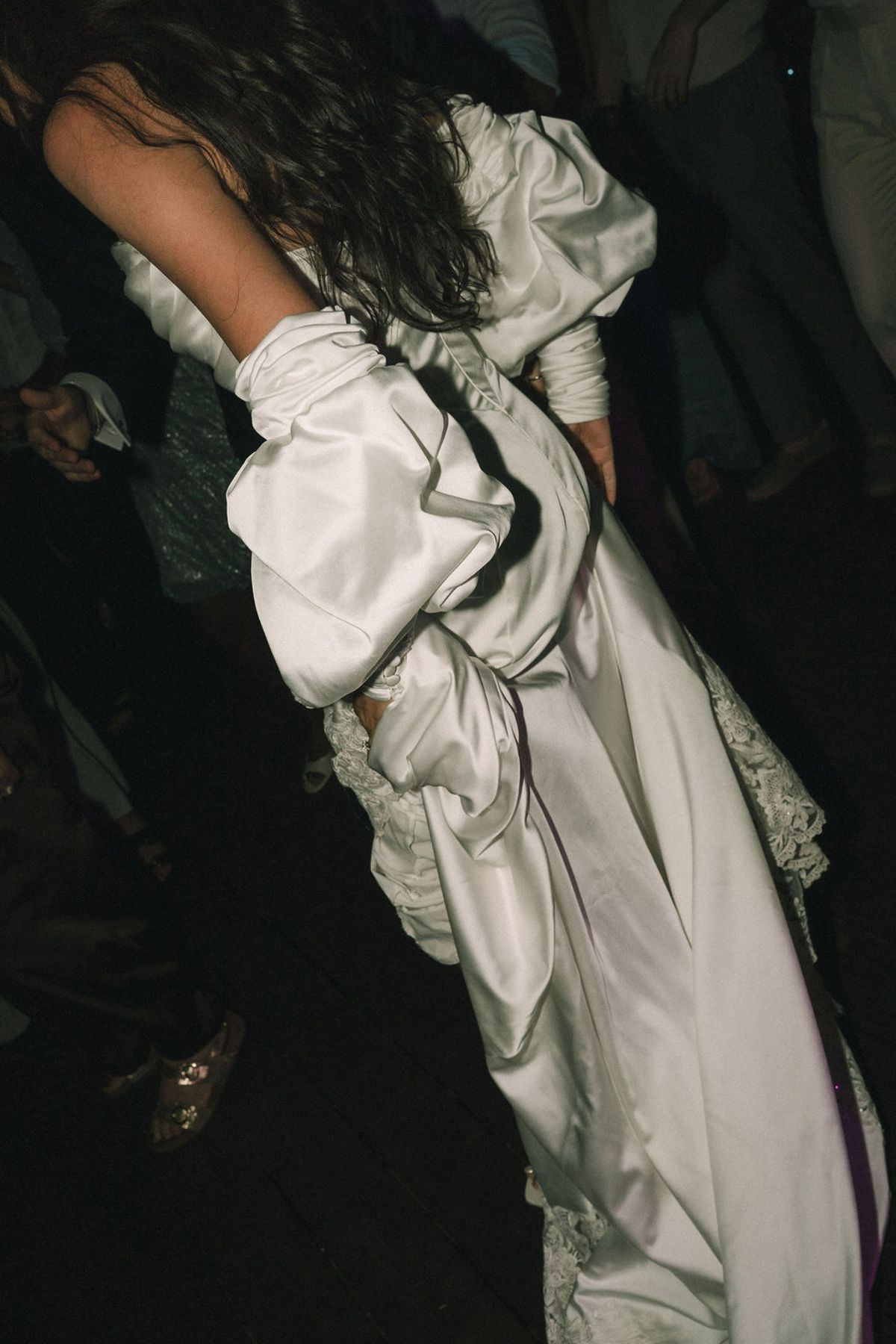 The bride, seen from behind in a dark room, wears a white satin gown with large puffed sleeves and long dark hair.