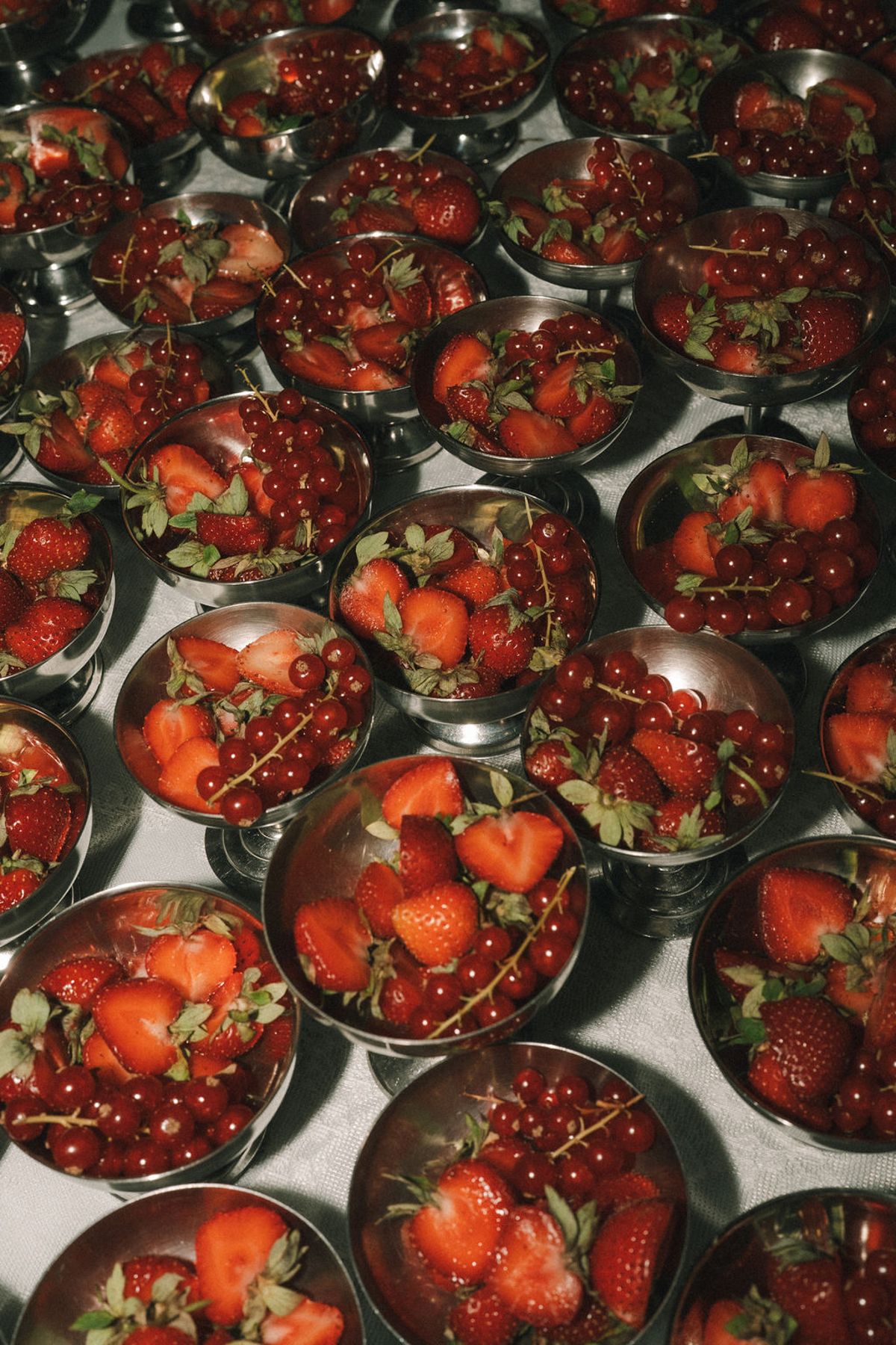 Many small metal bowls are filled with fresh strawberries and redcurrants, photographed closely from above.