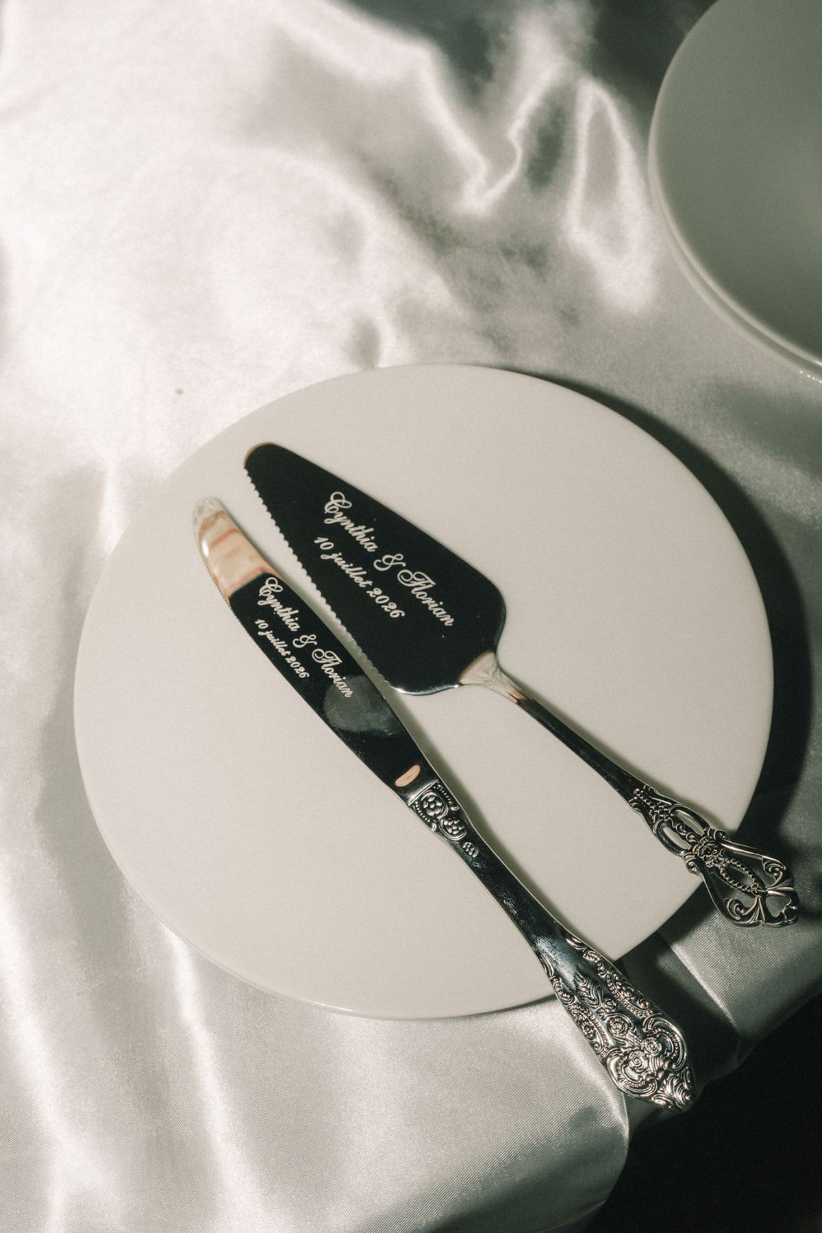 An engraved black cake knife and server lie on a white plate set on creased white satin, seen from above.