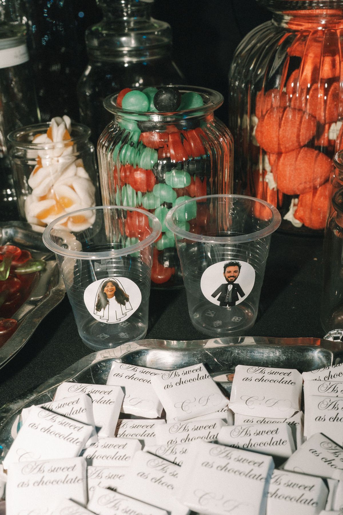 A dessert table with glass jars of colourful sweets, cups printed with small portraits and rows of chocolate notes.