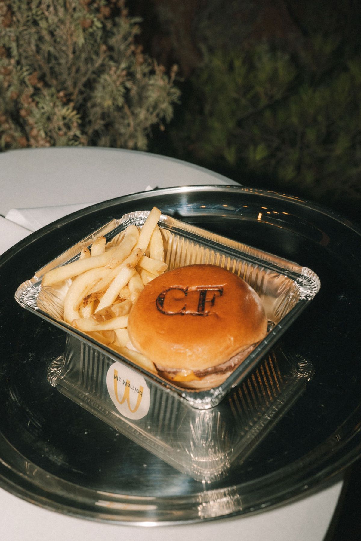 A burger with a monogram stamped on the bun and chips sit in a foil tray on a silver plate at night.
