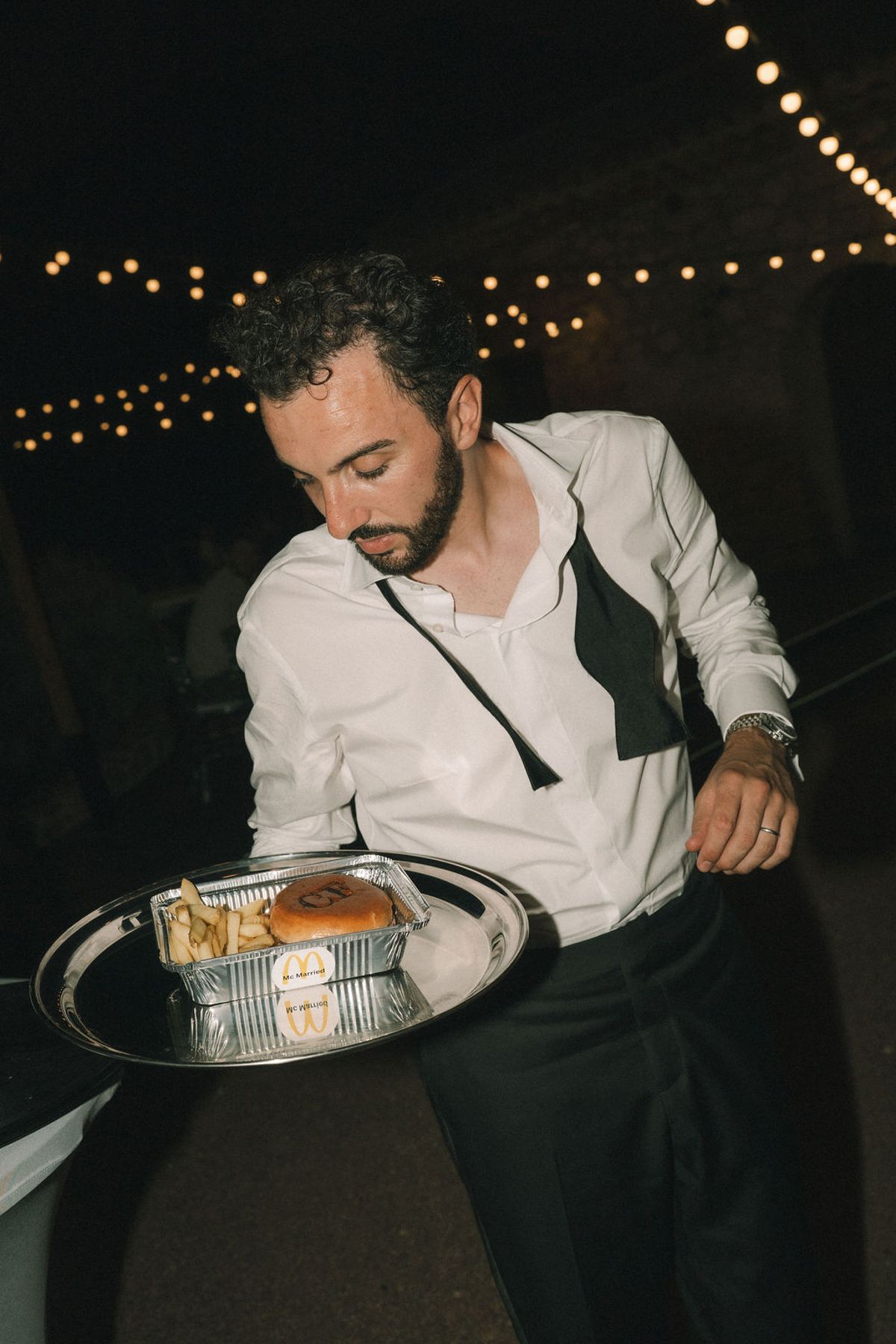 A bearded man in a white shirt with an undone bow tie carries a metal tray of food under string lights at night.