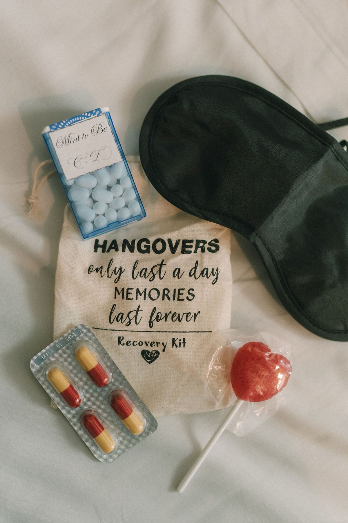 A recovery kit laid flat on white fabric includes a black sleep mask, mints, capsules, a red lollipop and a printed pouch.