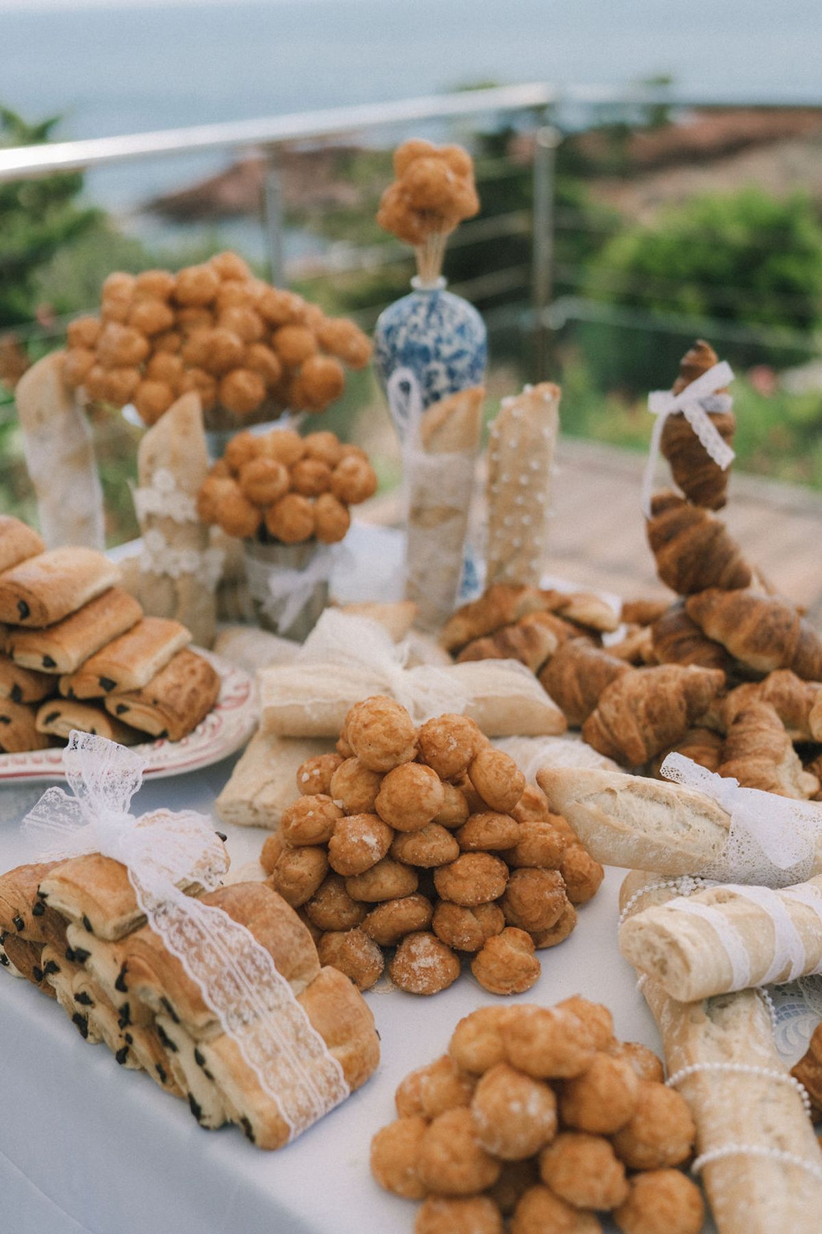 A table displays pastries wrapped in lace, piles of choux buns and a blue and white vase, with the sea beyond.