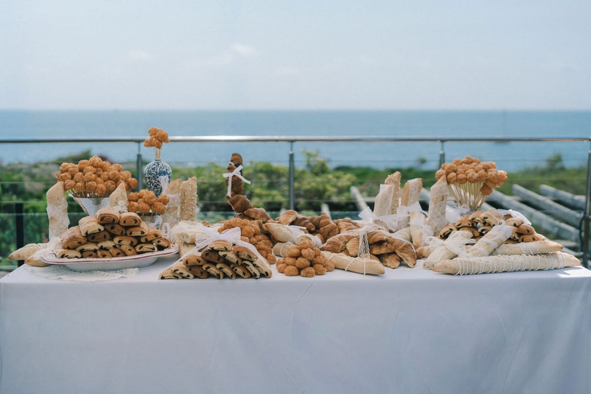 A long white-clothed table is covered with canapes and pastries on a terrace, with the sea and horizon behind.