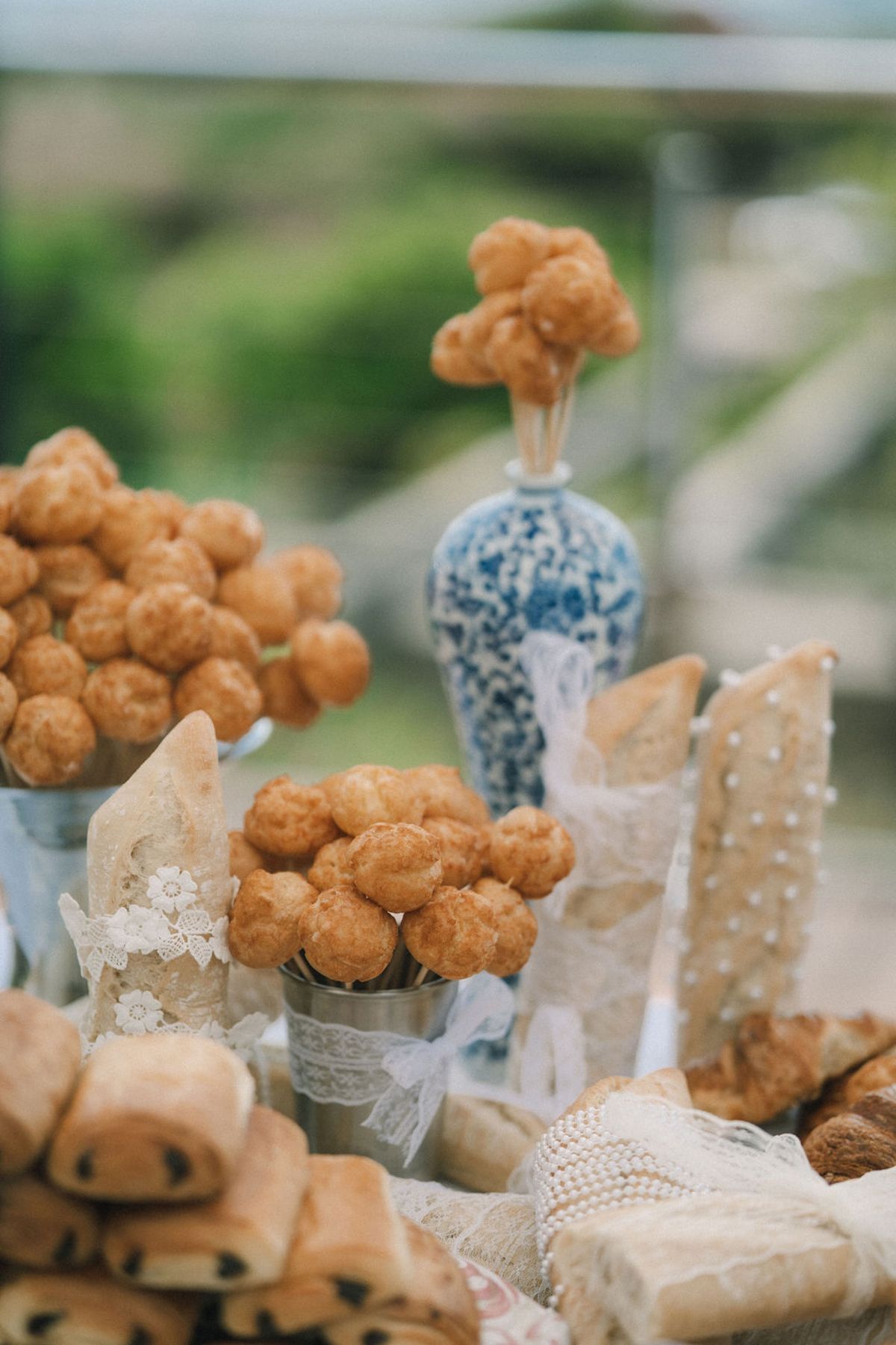 A display of golden choux buns, pastries and bread sits around a blue and white vase, with a blurred green background.