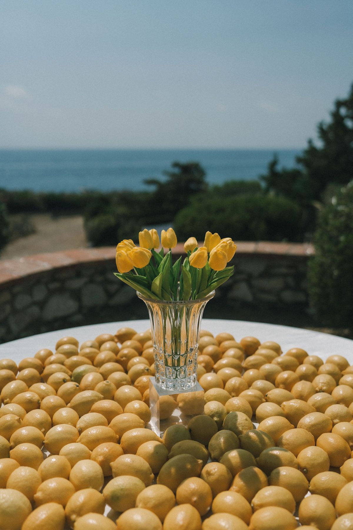 A glass vase of yellow tulips stands on a table covered in green olives, with the sea blurred behind.