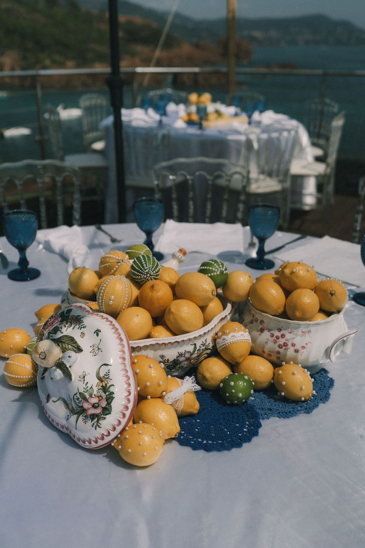 A white-clothed table holds a painted ceramic tureen and bowls of lemons, with blue glasses and a sea view behind.