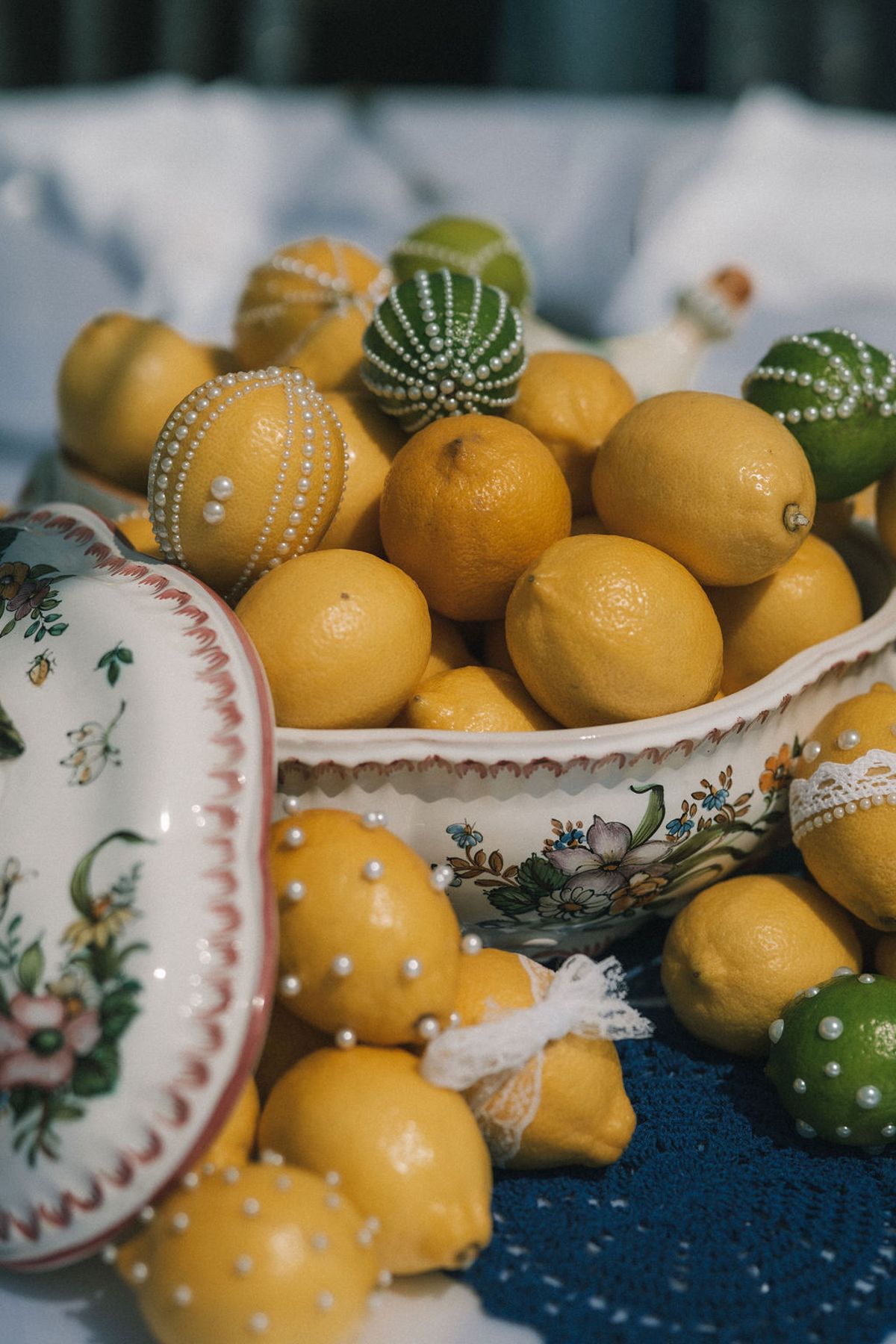 Close view of bright lemons and decorated limes piled in a floral porcelain bowl, resting on a blue crocheted mat.