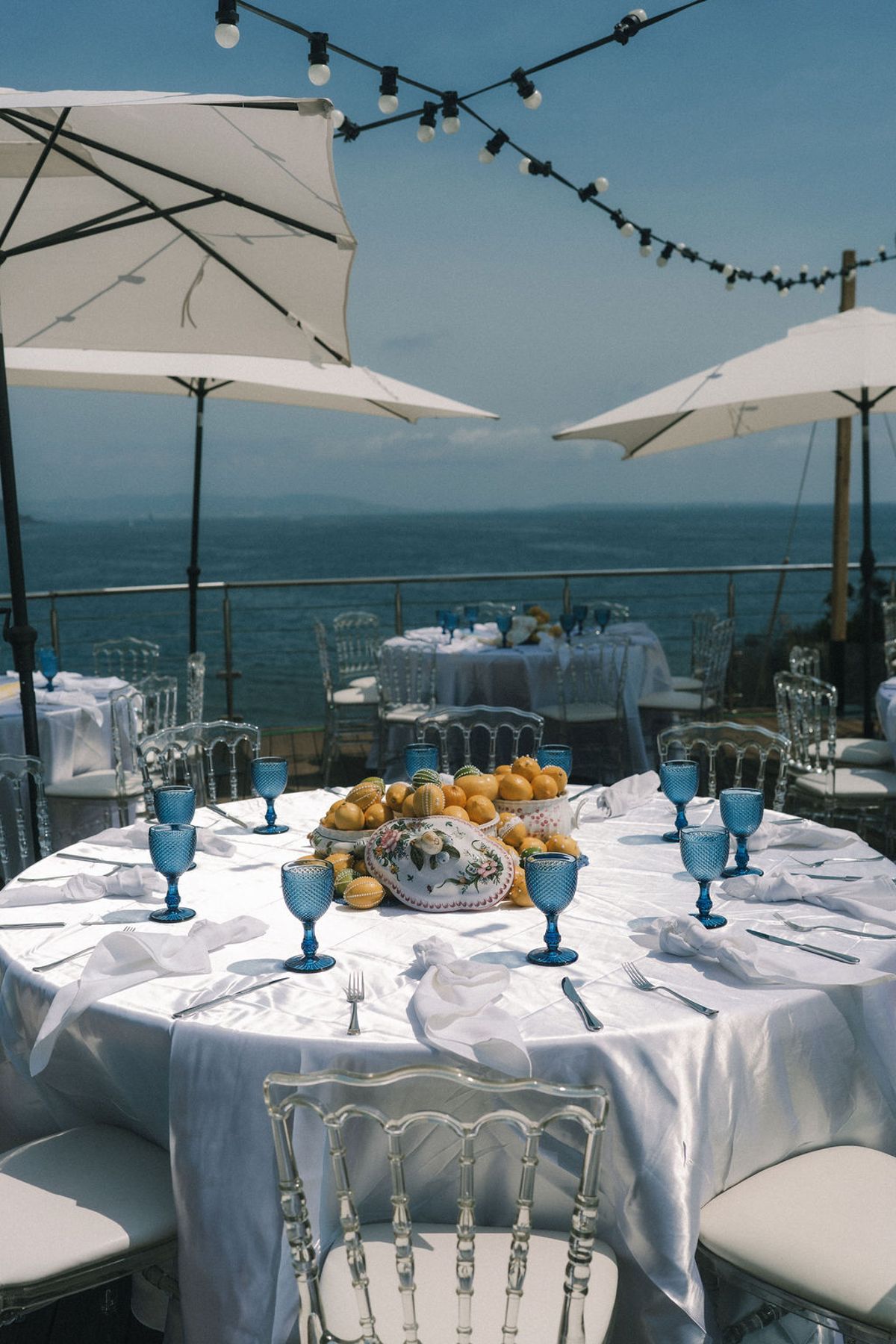 A round table with a white cloth, blue glasses and a bread basket stands on a terrace beneath parasols, with the sea behind.
