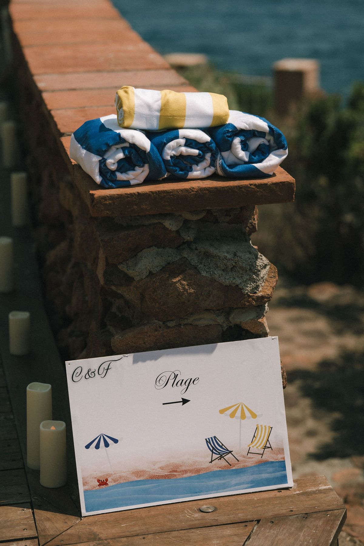 Rolled blue and yellow towels sit on a stone wall above an illustrated beach sign with an arrow, beside candles.