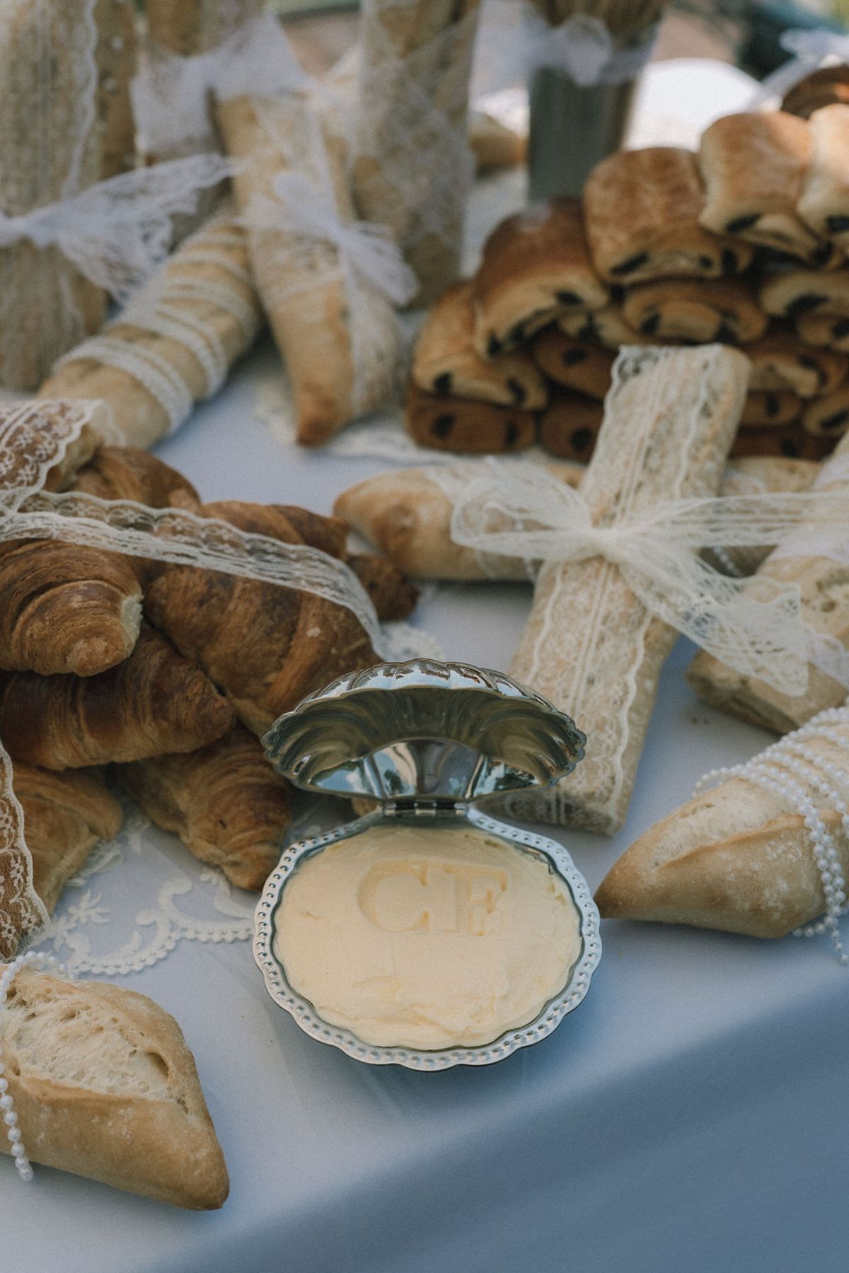 Croissants, pains au chocolat and baguettes tied with lace sit on a white cloth beside a shell dish of butter.