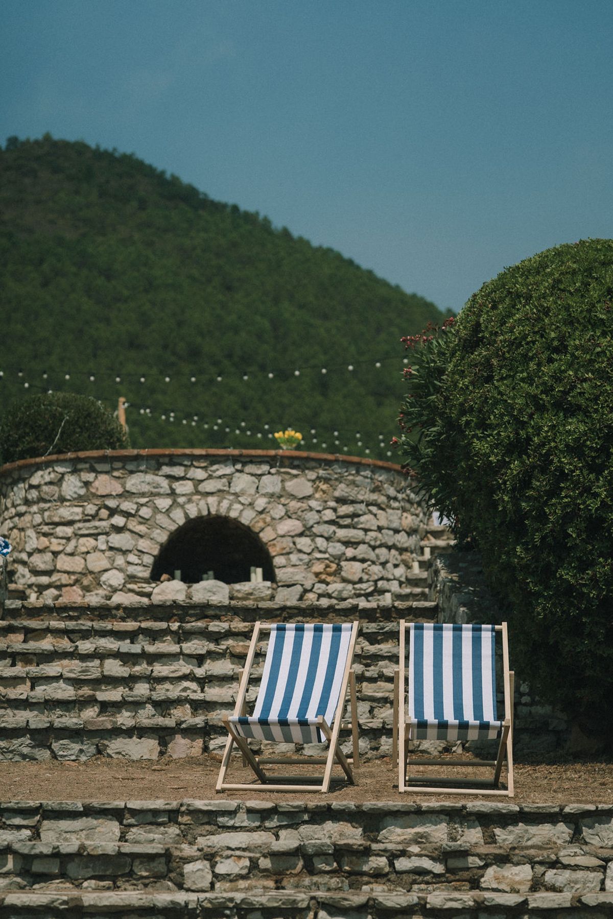 Two blue and white striped deckchairs stand on stone steps in front of a curved stone wall, with a wooded hill behind.