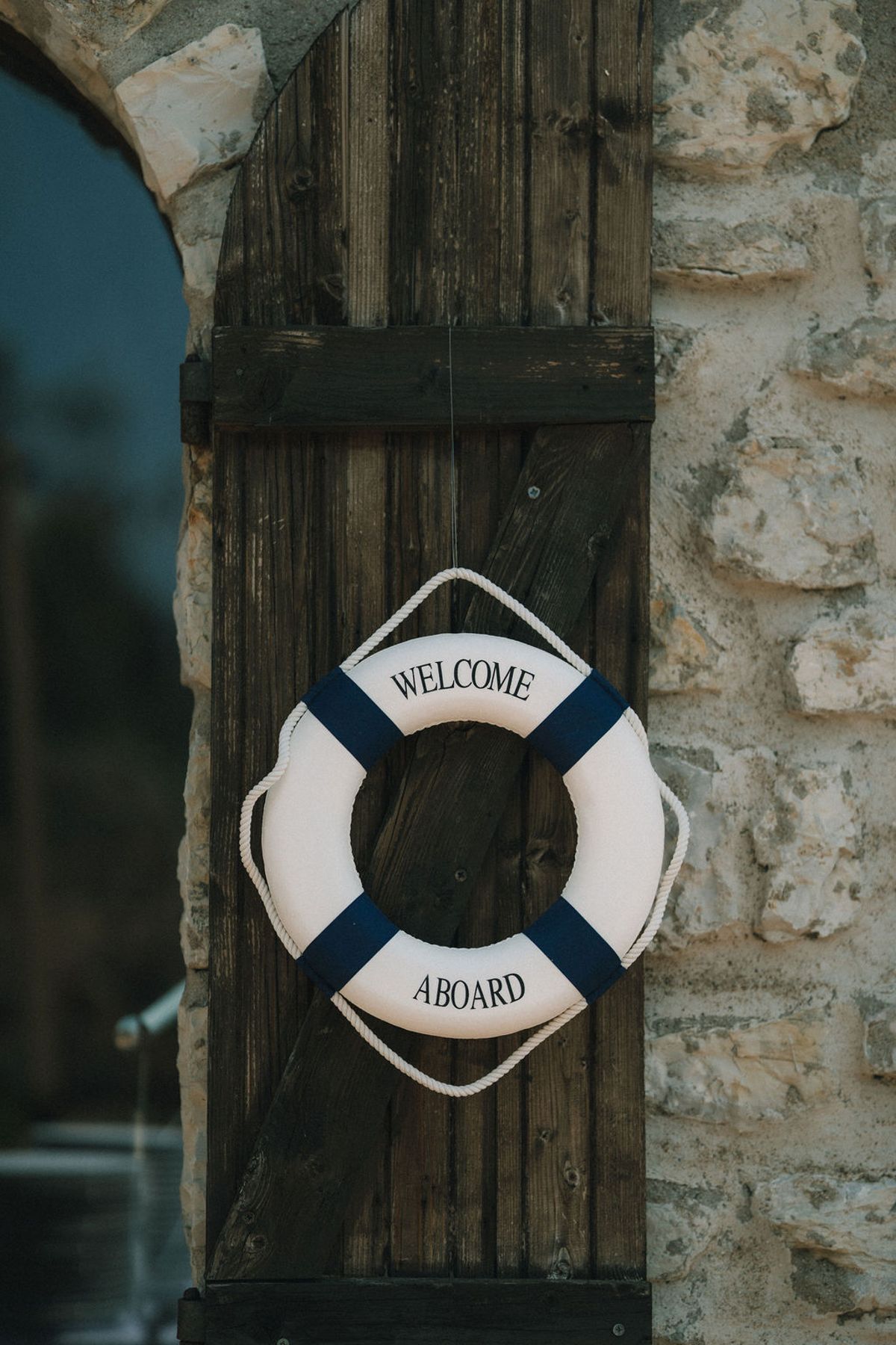 A white and navy lifebuoy reading Welcome Aboard hangs on a weathered wooden door beside a stone wall.