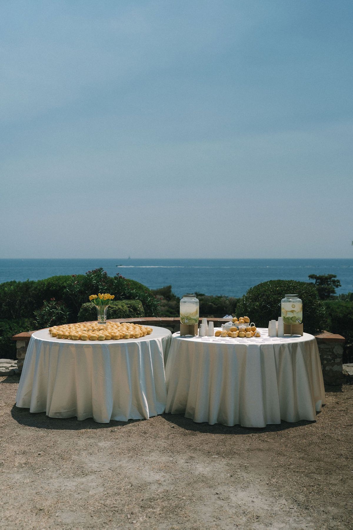 Two round tables with white cloths hold food and drink dispensers, set against a wide blue sea and clear sky.