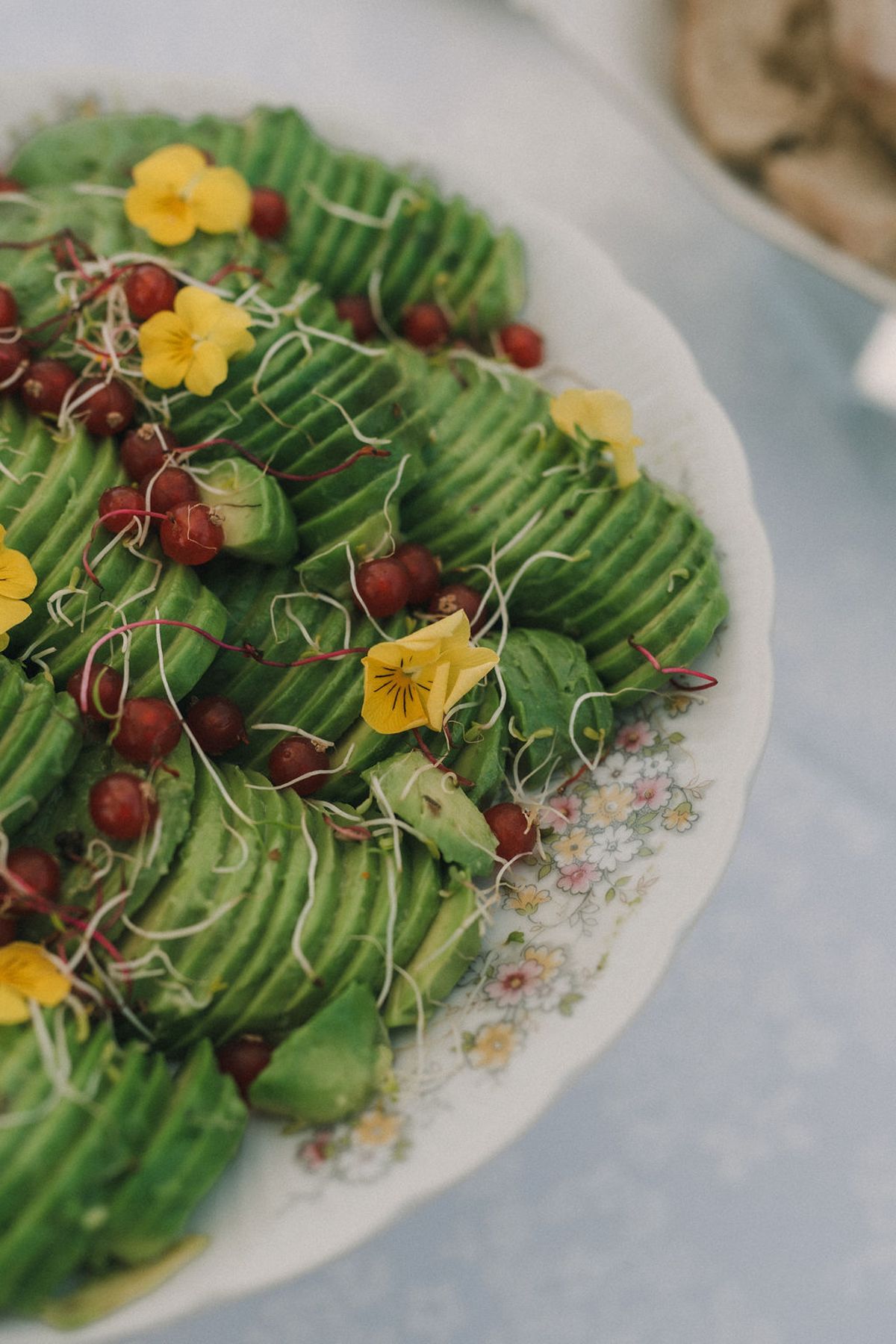 Sliced avocado topped with red berries and yellow edible flowers on a floral-rimmed plate on a white tablecloth.