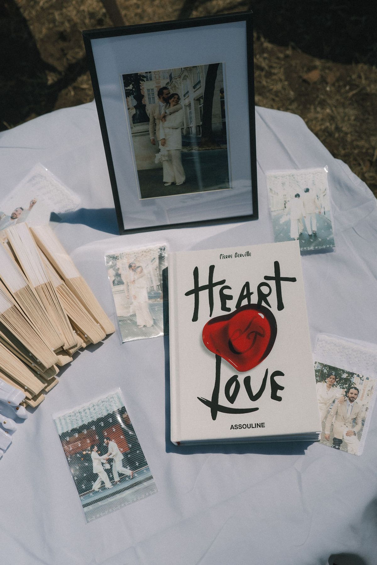 A round table holds a framed photograph, a book with a red heart on its cover, wooden fans and small photo cards.