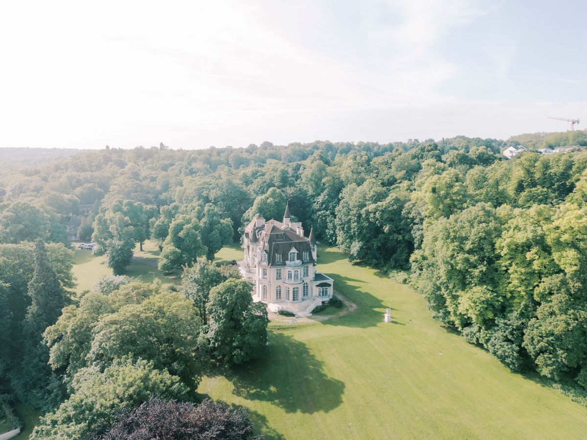 An aerial view shows a chateau surrounded by dense green woodland and a wide sweep of lawn.