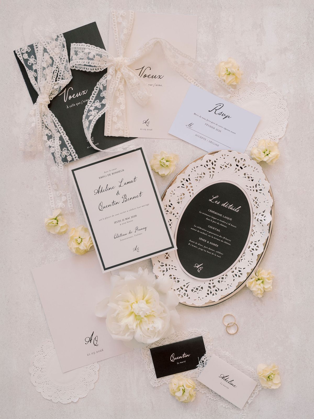 Black and cream invitations, menus and place cards are arranged with lace doilies, yellow flowers and two rings on a pale surface.