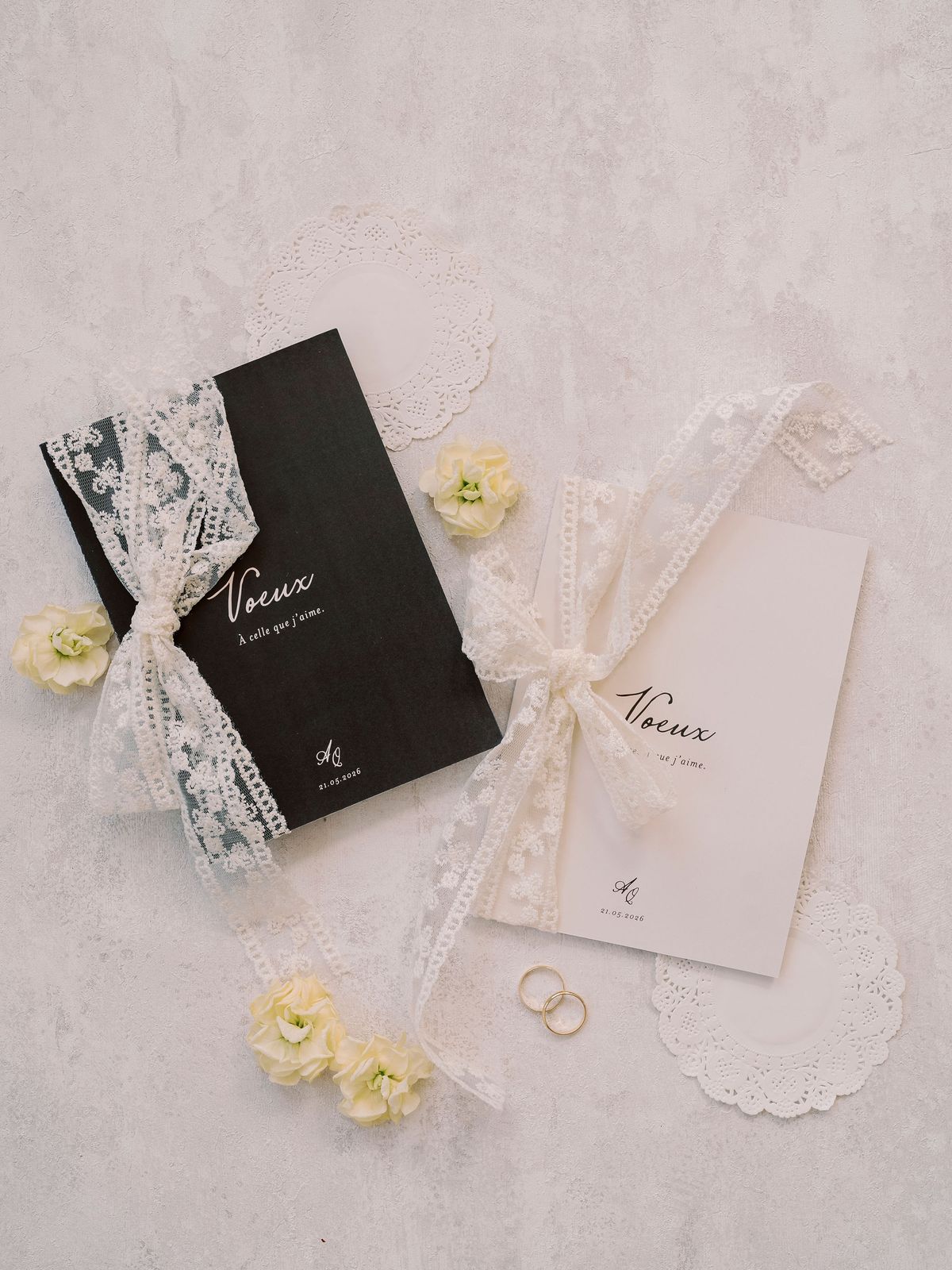Two vow booklets, one black and one cream, are tied with lace and laid with yellow flowers and two rings on a pale surface.