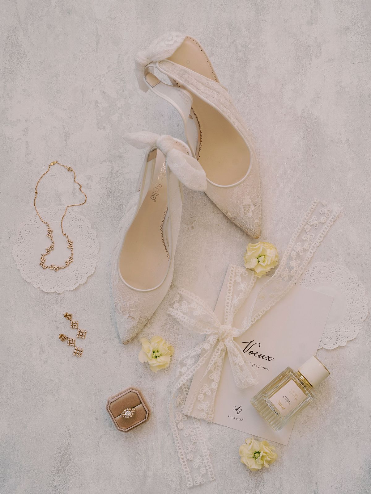 Cream bow-trimmed shoes, a necklace, a ring box, perfume and lace-tied stationery are laid on a pale surface with small yellow flowers.