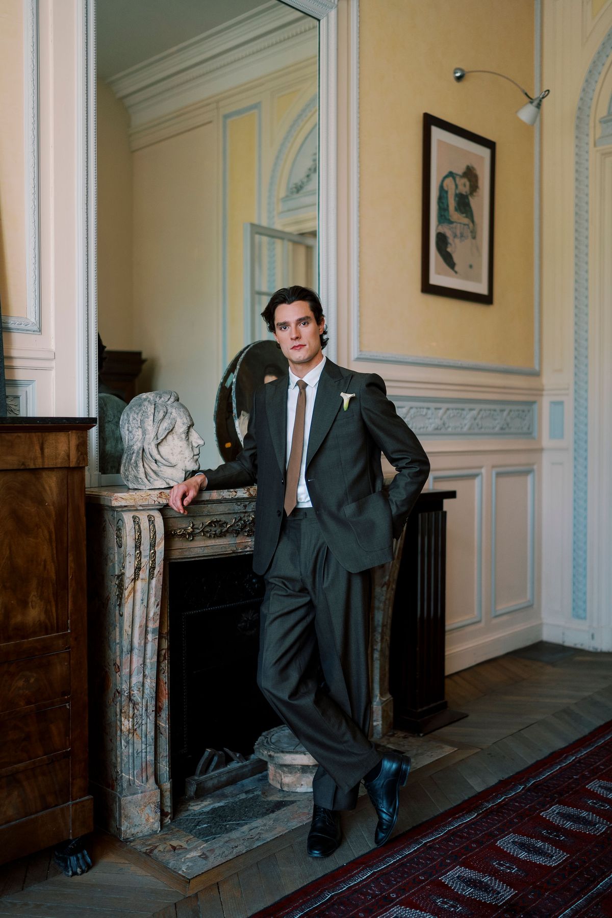 The groom in a grey suit leans casually against a marble fireplace in a yellow-walled room with a patterned rug.
