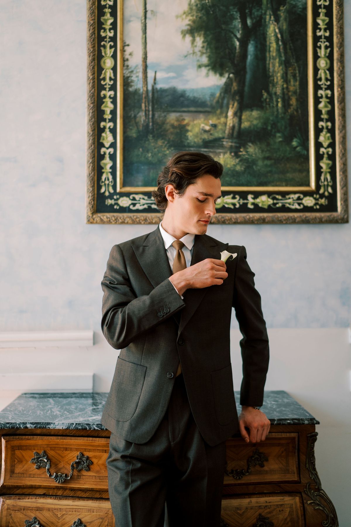 The groom in a dark grey suit adjusts a small flower on his lapel, leaning on a wooden chest beneath a framed landscape painting.