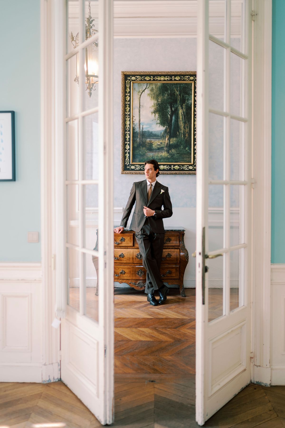 The groom in a dark suit stands in open white double doors, in a room with pale blue walls and a framed painting.