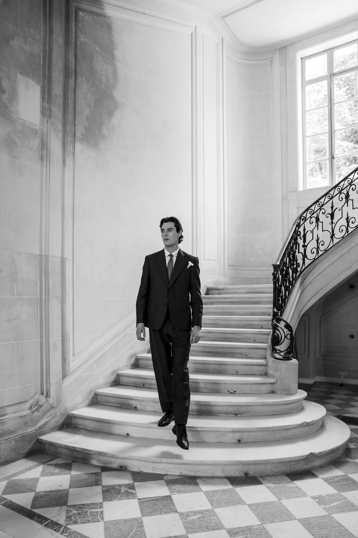 A man in a dark suit walks down a curved stone staircase with an ornate wrought-iron banister, in black and white.