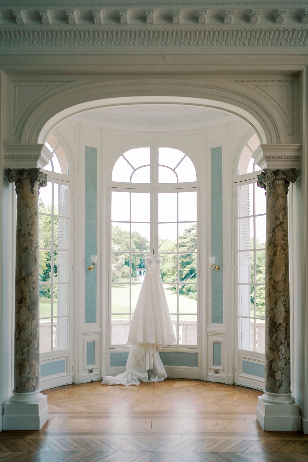 A white wedding gown hangs in front of tall arched windows between two marble columns, with a green park beyond.