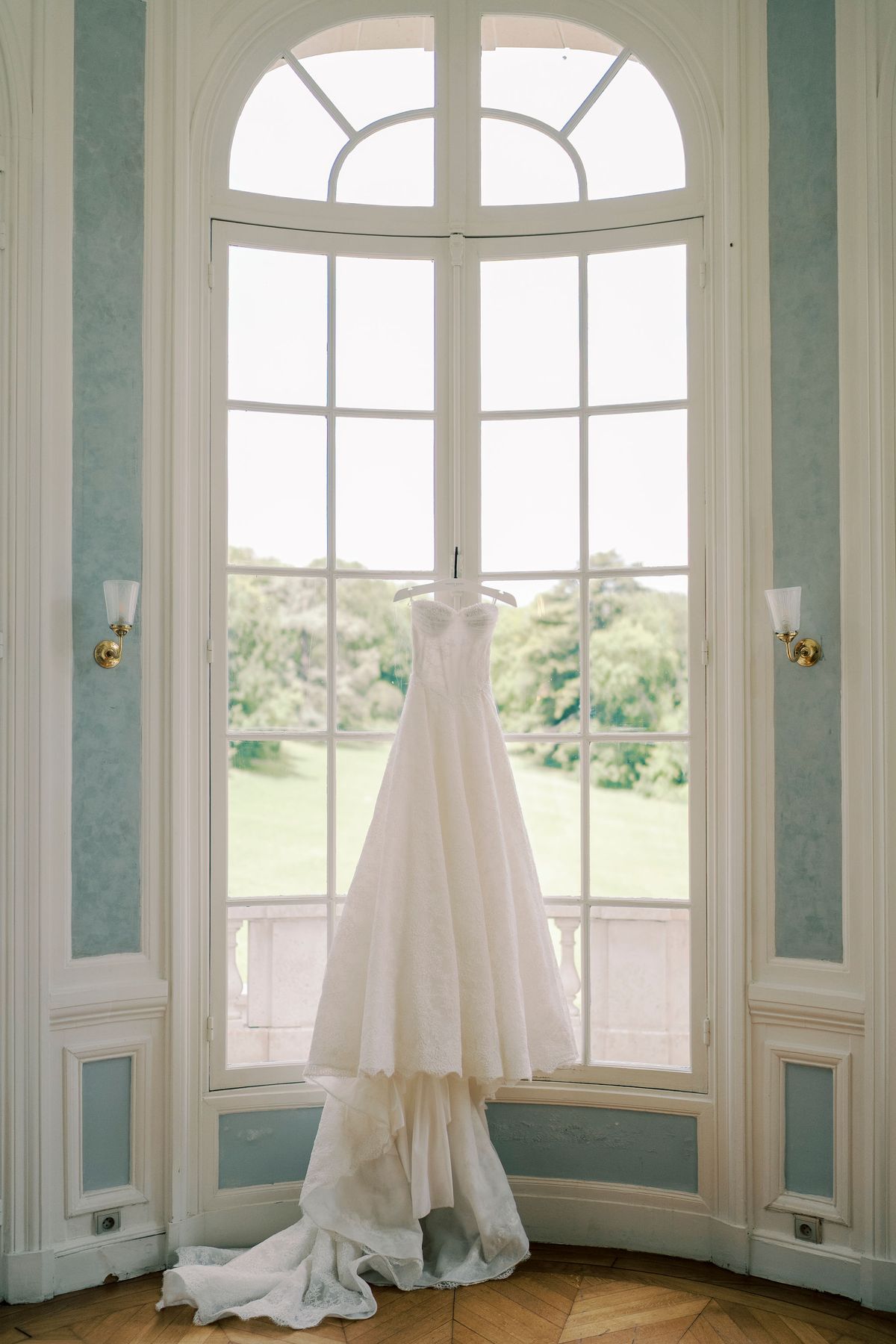 A white wedding dress with a long train hangs before a tall arched window with pale blue panelled walls and parkland beyond.