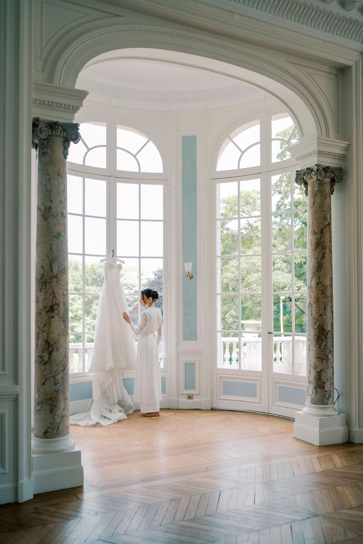 A wedding dress hangs in a bright rotunda with marble columns and tall arched windows, while a person stands beside it.