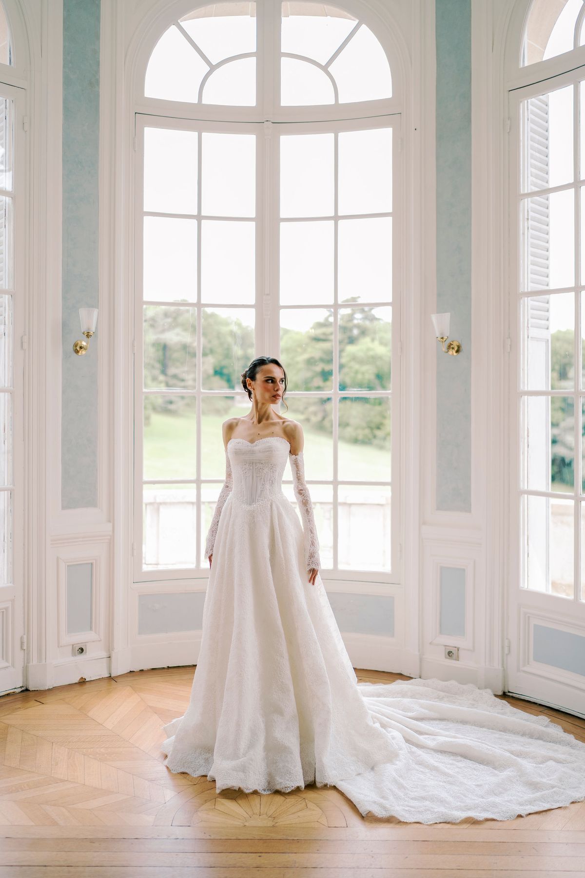 The bride in a white gown with a long train stands in a room with tall arched windows and a wooden parquet floor.