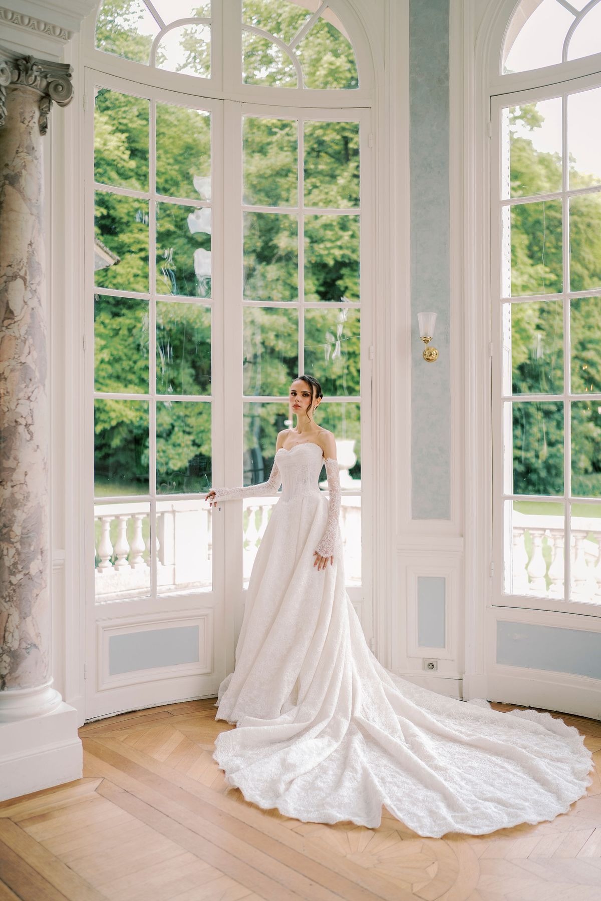 The bride stands in a curved window bay with tall glass doors, her long white gown train spread across the parquet floor.