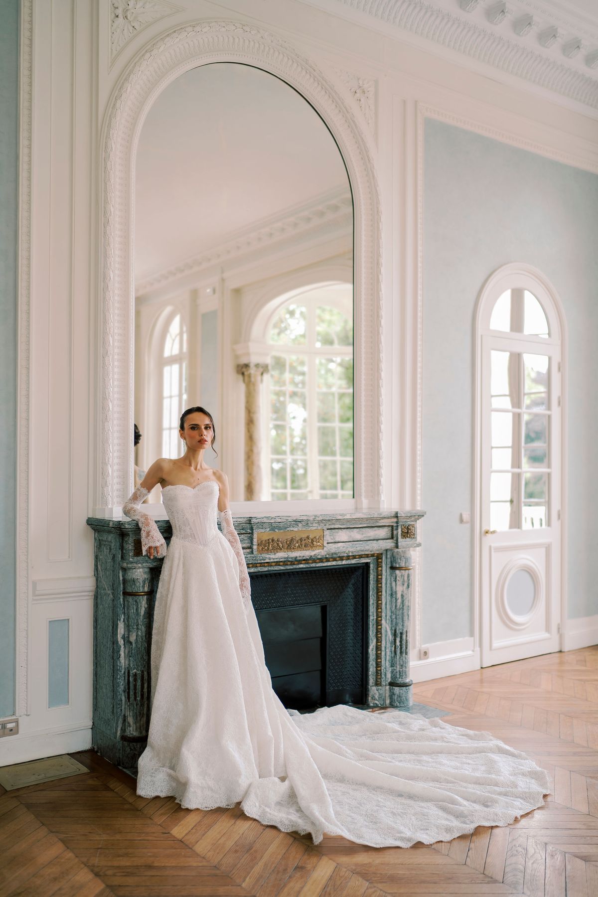 The bride stands beside a dark marble fireplace in a high-ceilinged room with pale blue panelling, her long gown trailing behind her.