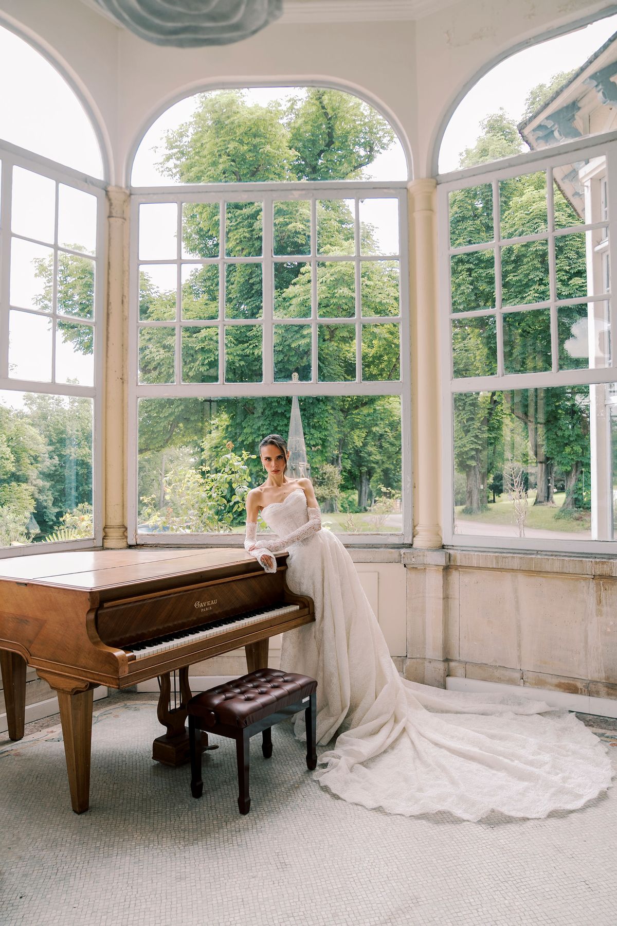 The bride sits at a wooden grand piano inside a rounded room with large arched windows looking onto trees, her gown spread around her.