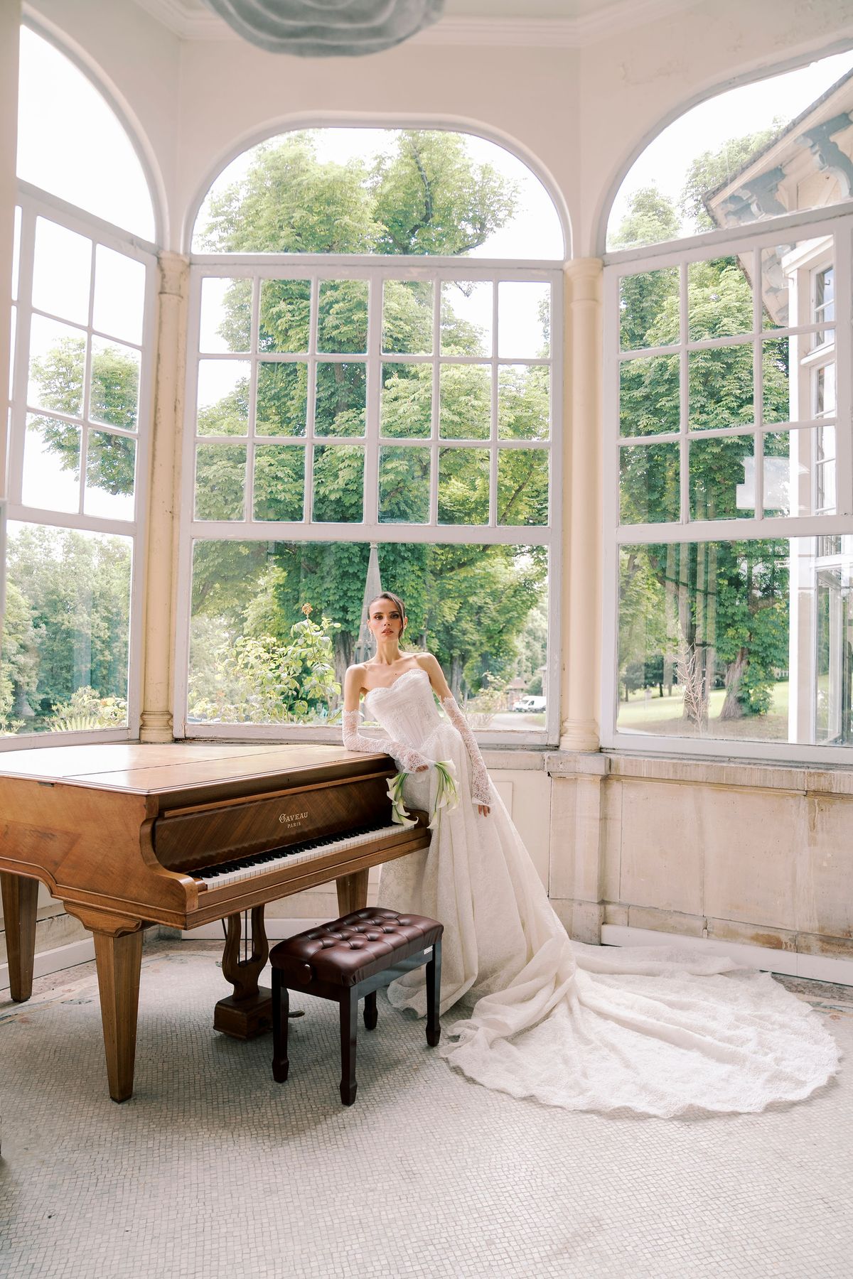 A bride in a long white gown sits beside a wooden grand piano in a bright room with arched windows overlooking trees.