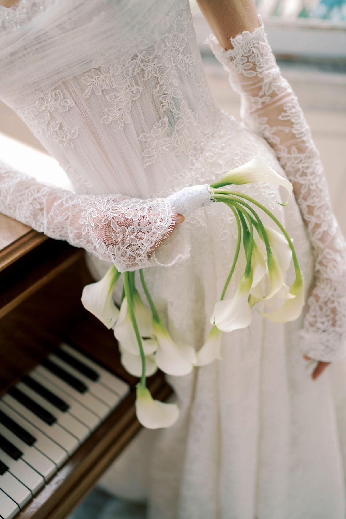 A bride in a lace gown and long lace gloves holds a bouquet of white calla lilies beside the keys of a piano.
