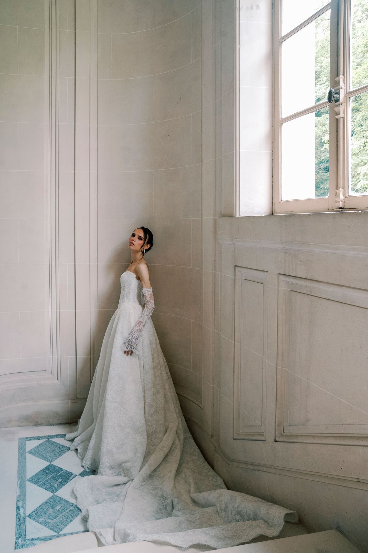 The bride in a white gown with long gloves and a full train stands against a pale panelled wall beside a tall window.