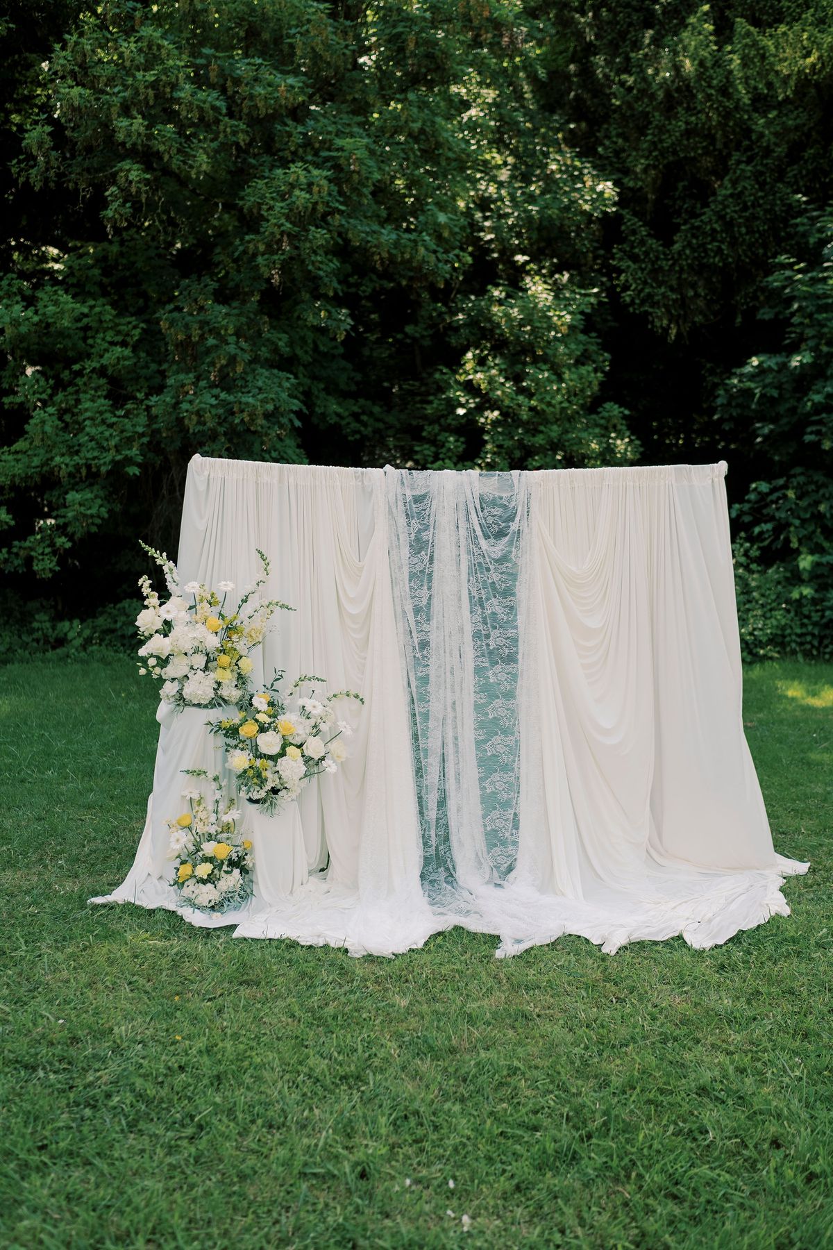 A backdrop of white draped fabric with a pale blue lace panel and yellow and white flowers stands on grass in front of dark trees.