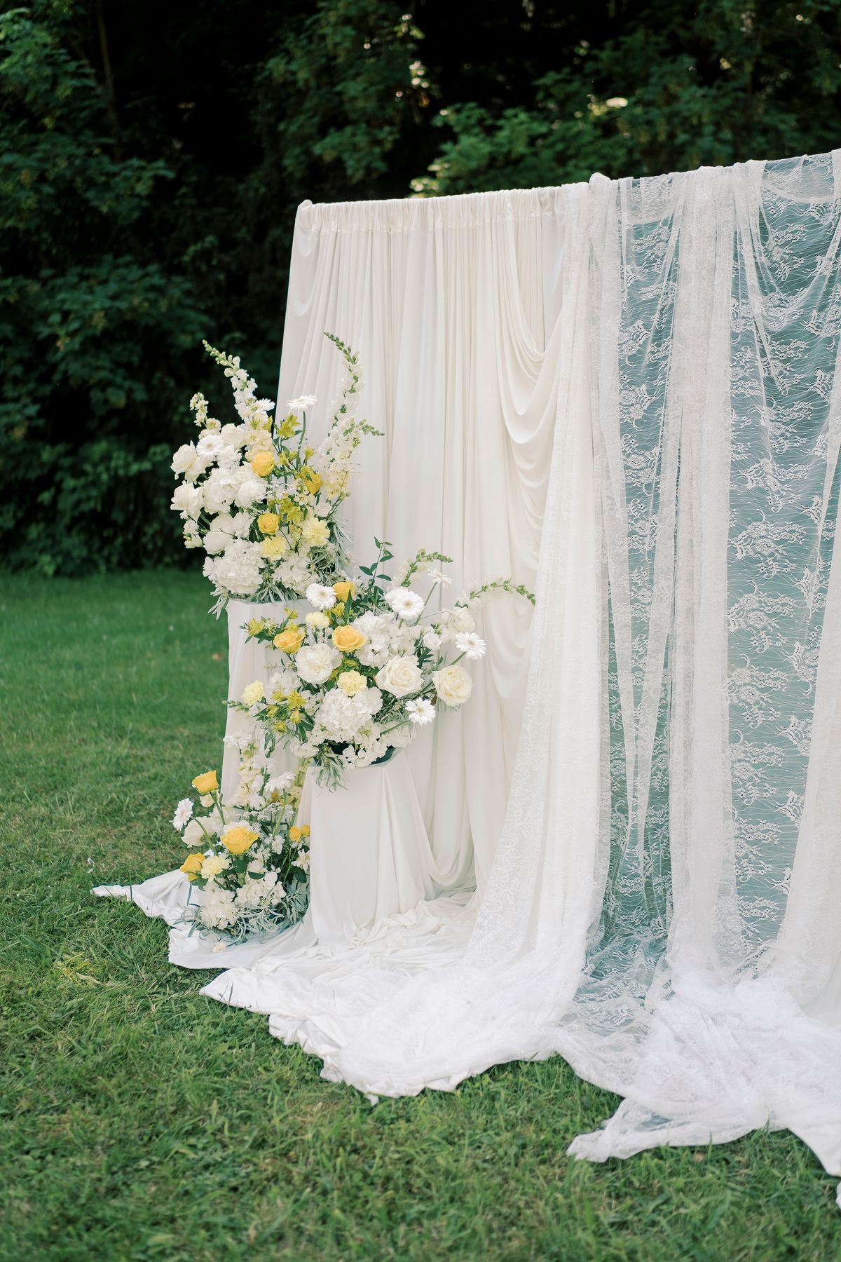 White draped fabric and a pale blue lace panel hang as a backdrop on a lawn, with yellow and white flowers at its base.